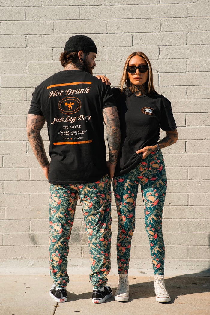 A man and woman pose before a light brick wall in black tees; she wears Tango Charlie Apparel's Women's Performance Leggings | Aloha AR, featuring a floral print and tummy control, while he shows off his shirt’s back print. She touches his shoulder, wearing sunglasses.