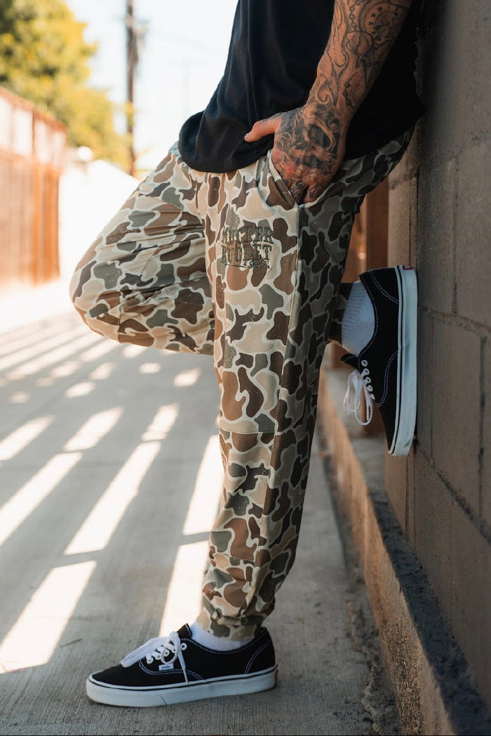 A tattooed person leans against a wall outdoors, wearing Mucker Bucket Men's Classic Premium Joggers in Duck Camo and black Vans. Sunlight filters through a fence, casting shadows on the sidewalk. The person's face is hidden.