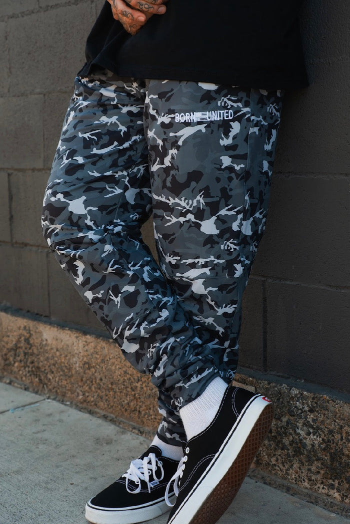 A person stands against a dark brick wall with one foot crossed, wearing Born United Private Label Men's Classic Premium Joggers in Gunmetal Camo, black Vans sneakers with white laces, and white socks.