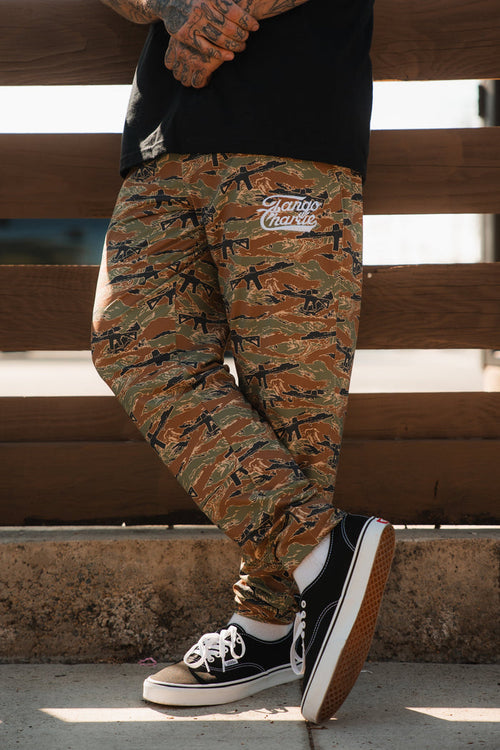 A person stands outdoors wearing Tango Charlie Apparel’s Men's Classic Premium Joggers | Snipes & Stripes—camouflage pants with Hando Master text—and black-and-white sneakers. Only the lower half shows, set against wooden fencing. Quick-dry fabric suits running.
