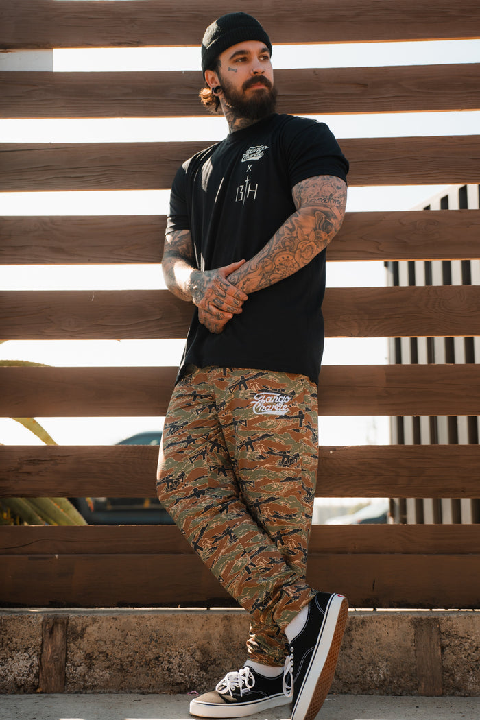 A tattooed man in a black beanie and t-shirt stands by a wooden fence, sporting Tango Charlie Apparel’s Men’s Classic Premium Joggers | Snipes & Stripes, camo print with quick-dry fabric, paired with black sneakers.