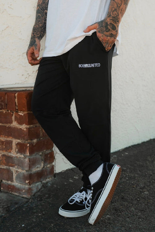 A person with tattooed arms, dressed in a white t-shirt, black Vans, and Born United Private Label Men's Classic Premium Joggers | Stealth, stands casually against a brick ledge and white wall.