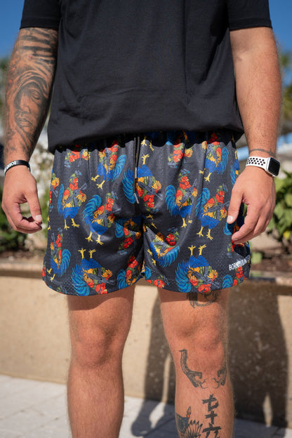 A tattooed person outdoors wears a black shirt and Born United Private Label Men's Mesh Lifestyle Shorts | 5.5" - Battle Bird, featuring a colorful rooster pattern, along with a watch and wristband.