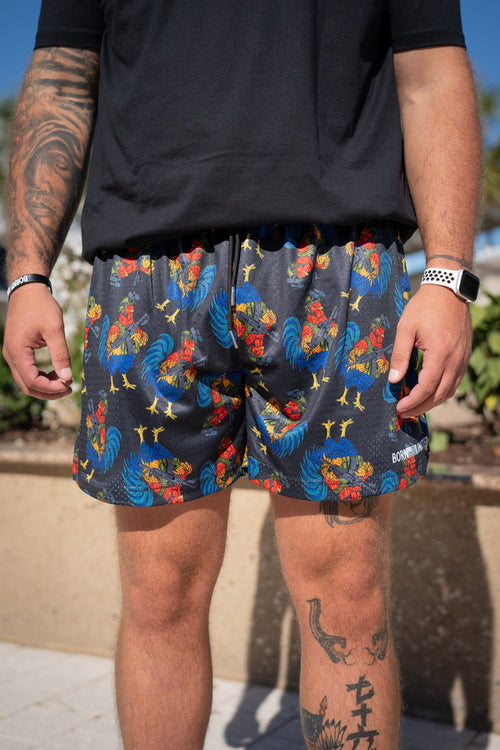 A tattooed person outdoors wears a black shirt and Born United Private Label Men's Mesh Lifestyle Shorts | 5.5" - Battle Bird, featuring a colorful rooster pattern, along with a watch and wristband.