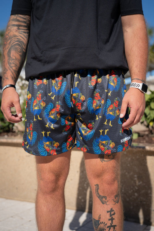 A tattooed person outdoors wears a black shirt and Born United Private Label Men's Mesh Lifestyle Shorts | 5.5" - Battle Bird, featuring a colorful rooster pattern, along with a watch and wristband.