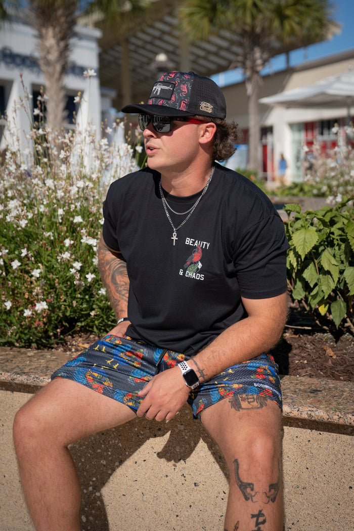 A man in a black cap, black Beauty & Chaos T-shirt, sunglasses, and colorful Born United Private Label Men's Mesh Lifestyle Shorts | 5.5"- Battle Bird sits outdoors on a low wall amid flowers and greenery with buildings and palm trees behind him.