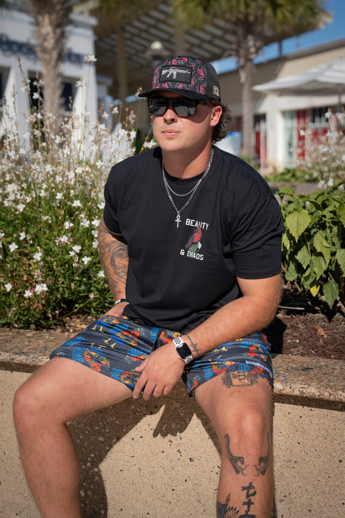 A man in a black t-shirt, sunglasses, and cap sits on a low wall outdoors wearing Born United Private Label Men's Mesh Lifestyle Shorts | 5.5" - Battle Bird. He has arm and leg tattoos; plants and buildings are visible in the background.