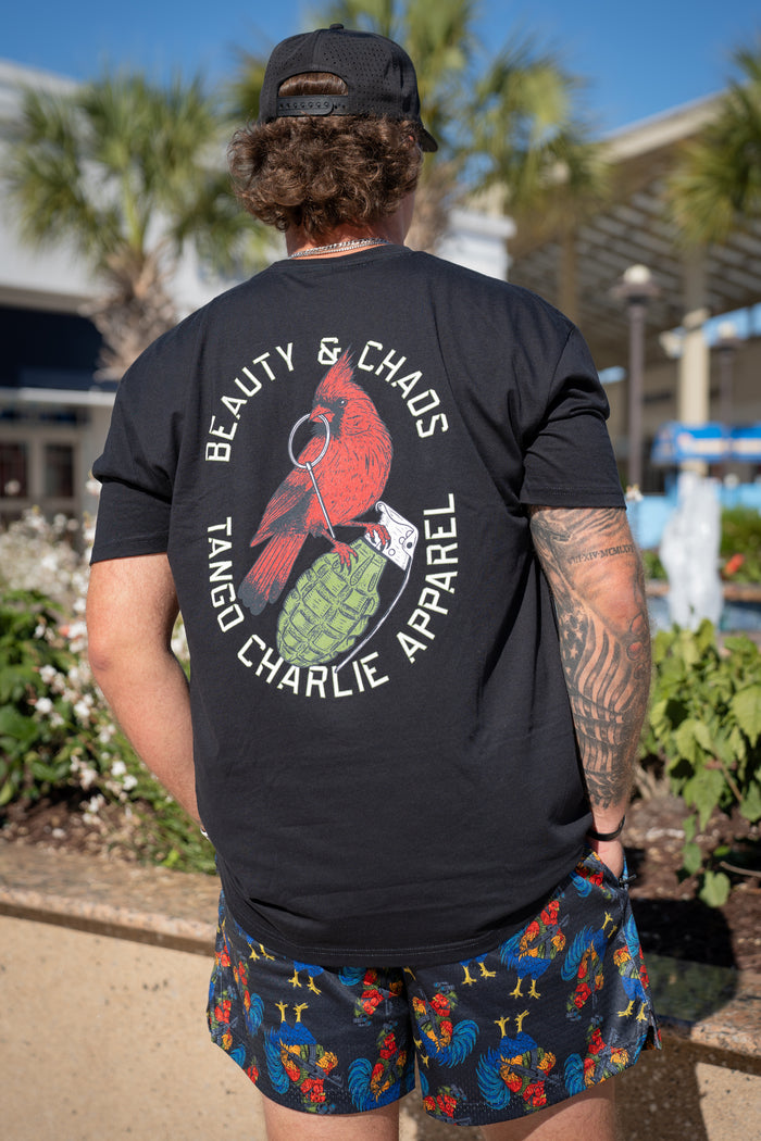 Wearing Born United Private Label Men's Mesh Lifestyle Shorts | 5.5" - Battle Bird, a person with curly hair, tattoos, a black cap, and graphic tee stands outdoors among greenery and palm trees, facing away from the camera.