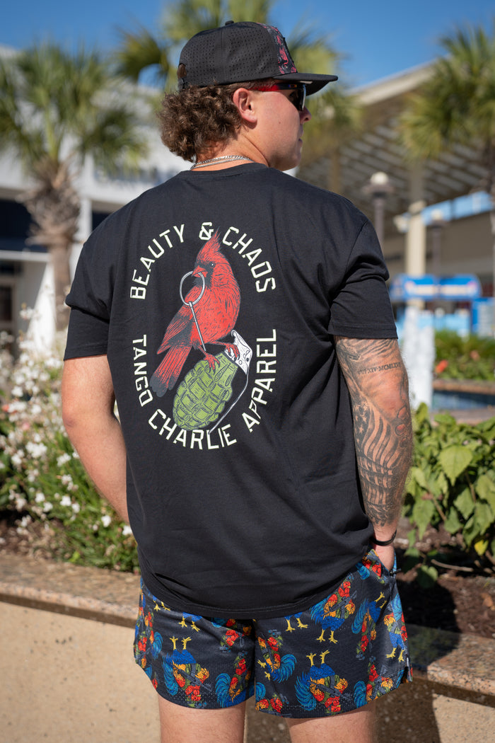 A tattooed person with curly hair wears a black cap, a black tee with a red bird and grenade graphic, and Born United Private Label Men's Mesh Lifestyle Shorts | 5.5"- Battle Bird, standing outdoors near greenery under the sunny sky.