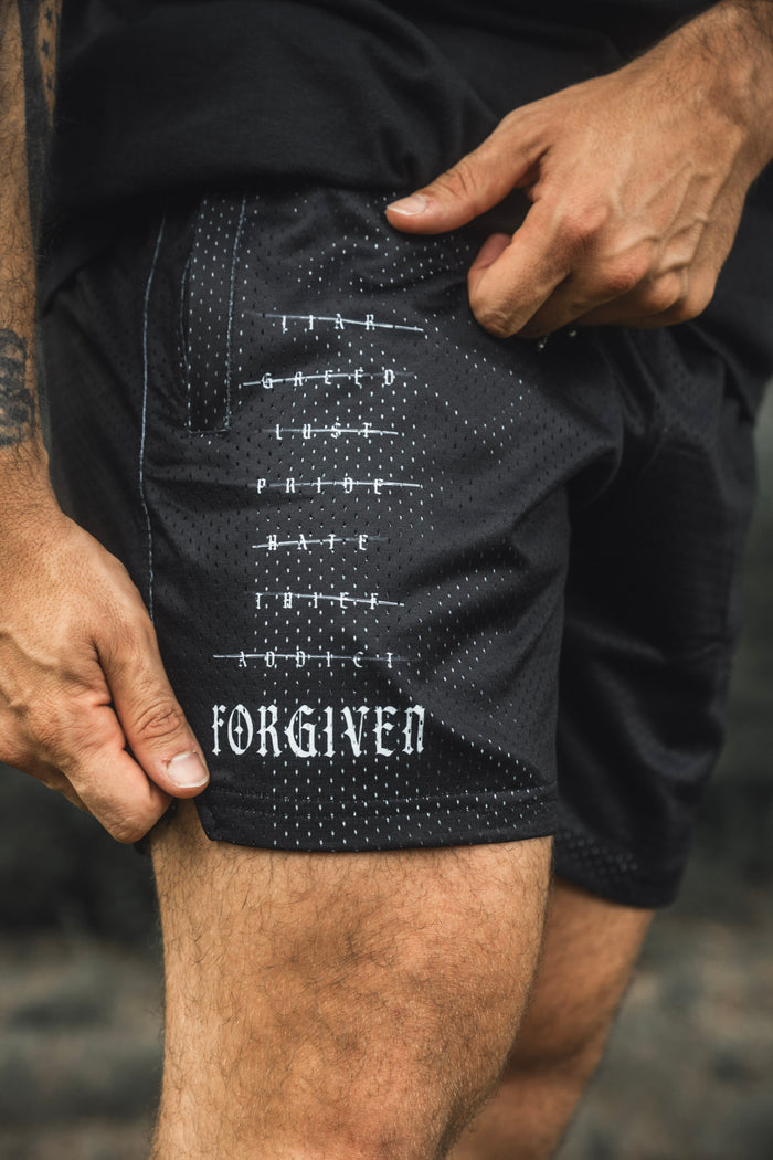 Wearing the 13th Men’s Mesh Lifestyle Shorts | 5.5″- Forgiven, a person sports black unisex mesh shorts crafted from breathable performance material. The design features the words Fear, Greed, and Hate crossed out, with “Forgiven” boldly displayed on the left leg.