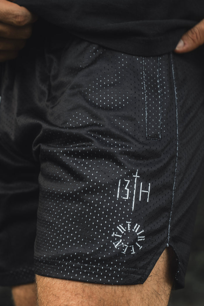 A person is wearing the Men’s Mesh Lifestyle Shorts | 5.5″ - Forgiven by 13th, featuring a black mesh fabric with a white dotted pattern, convenient pocket, and distinctive graphics—including “13TH” and a clock motif on the left leg. These unisex shorts combine comfort and style, perfect for any active lifestyle.