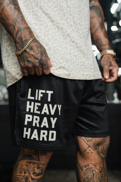 A person with tattooed arms and legs wears Tango Charlie Apparel Men's Mesh Lifestyle Shorts | 5.5" - Lift Heavy Pray Hard Black, featuring "LIFT HEAVY PRAY HARD" in bold white text, standing indoors.