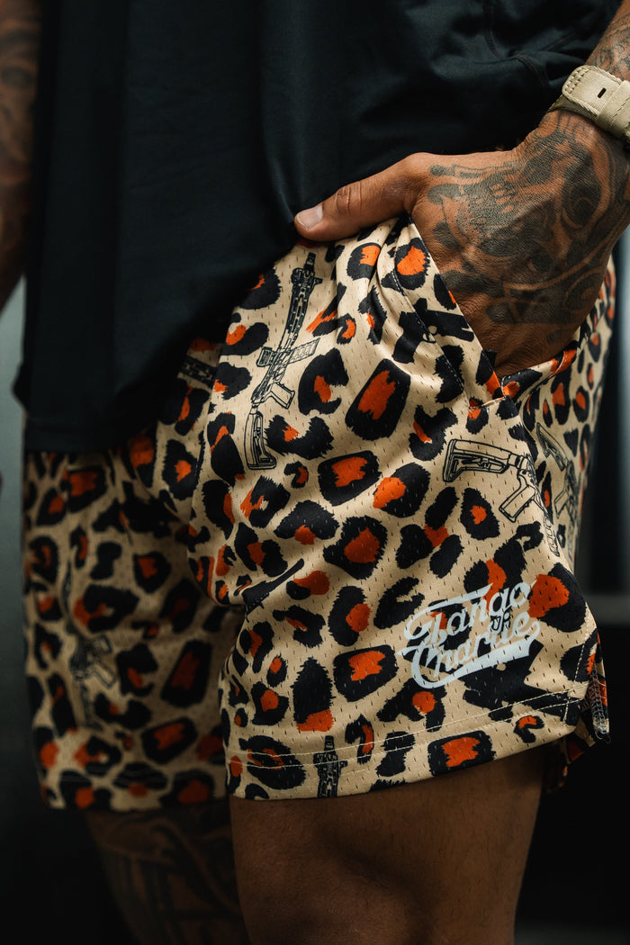 A person with tattooed arms wears a black shirt and Tango Charlie Apparel’s Men's Mesh Lifestyle Shorts (5.5" Loaded Leopard) featuring a leopard print, white logo, and handguns, paired with a beige wristwatch and one hand in their pocket.