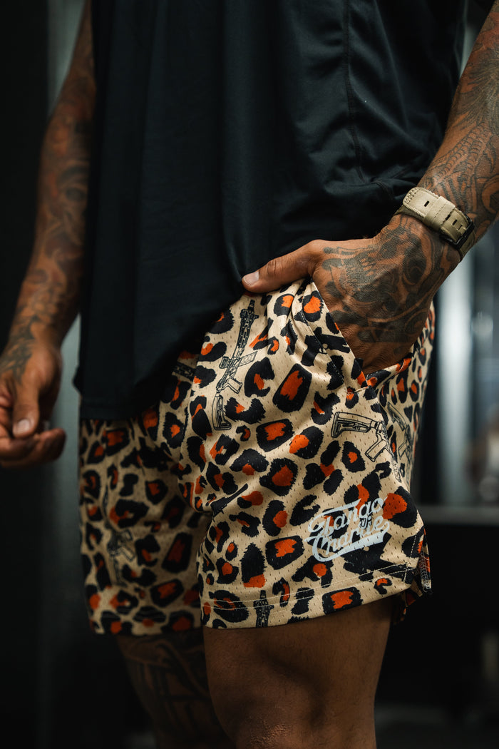 A person with tattooed arms, hand in pocket, wears a black shirt and Tango Charlie Apparel Men's Mesh Lifestyle Shorts | 5.5"- Loaded Leopard, with the “Chango” logo visible on the shorts.