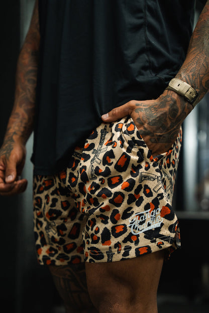 A person with tattooed arms, hand in pocket, wears a black shirt and Tango Charlie Apparel Men's Mesh Lifestyle Shorts | 5.5"- Loaded Leopard, with the “Chango” logo visible on the shorts.