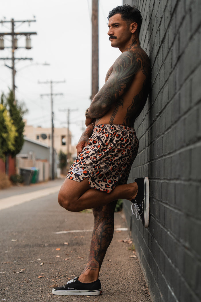 A shirtless, tattooed man leans against a dark brick wall outdoors, wearing Tango Charlie Apparel's Men's Mesh Lifestyle Shorts | 5.5"- Loaded Leopard and black sneakers, with one leg bent as he looks into the distance.