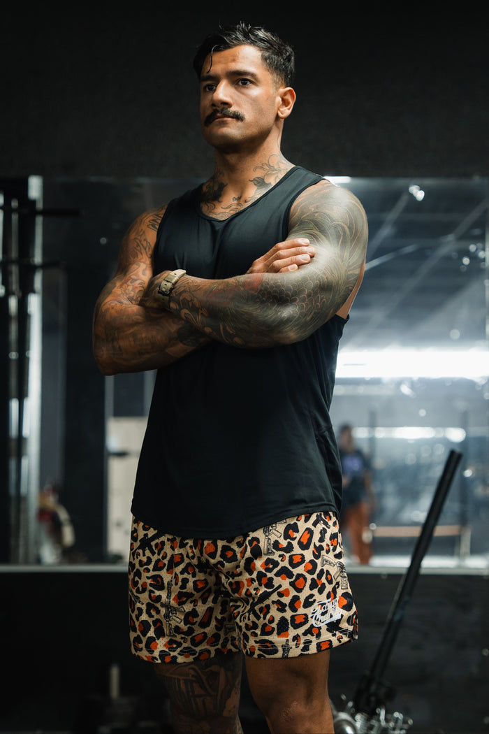 A muscular man with tattoos, wearing a black sleeveless shirt and Tango Charlie Apparel's Men's Mesh Lifestyle Shorts | 5.5"- Loaded Leopard, stands in a gym with his arms crossed, looking serious. Gym equipment is visible in the background.