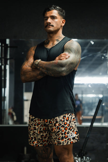 A muscular man with tattoos, wearing a black sleeveless shirt and Tango Charlie Apparel's Men's Mesh Lifestyle Shorts | 5.5"- Loaded Leopard, stands in a gym with his arms crossed, looking serious. Gym equipment is visible in the background.