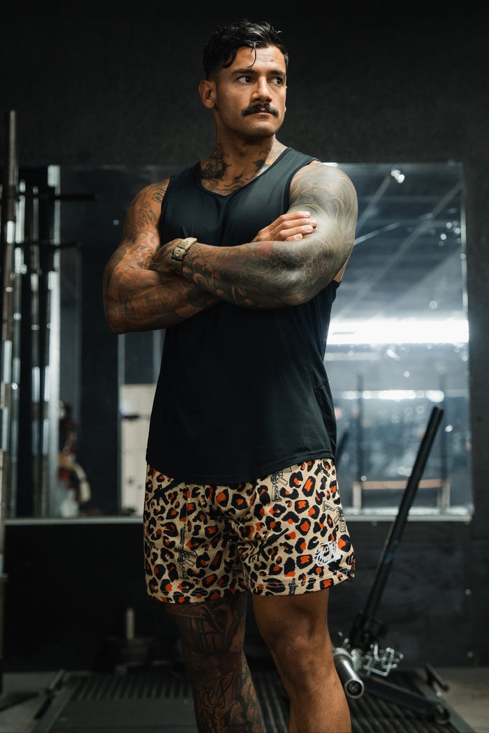 A muscular man with tattoos stands in a gym, arms crossed, wearing Tango Charlie Apparel’s Men's Mesh Lifestyle Shorts | 5.5"- Loaded Leopard and a black tank top. Gym equipment and a large mirror are visible in the background.