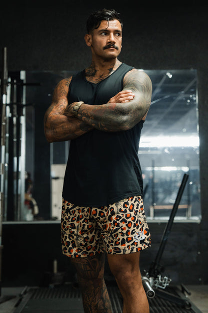 A muscular man with tattoos stands in a gym, arms crossed, wearing Tango Charlie Apparel’s Men's Mesh Lifestyle Shorts | 5.5"- Loaded Leopard and a black tank top. Gym equipment and a large mirror are visible in the background.