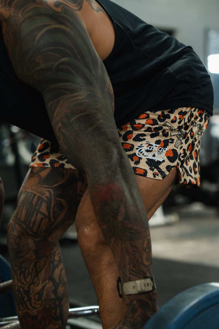A tattooed person in a black shirt and Tango Charlie Apparel Men's Mesh Lifestyle Shorts (5.5"- Loaded Leopard) bends over to lift a barbell in a gym.