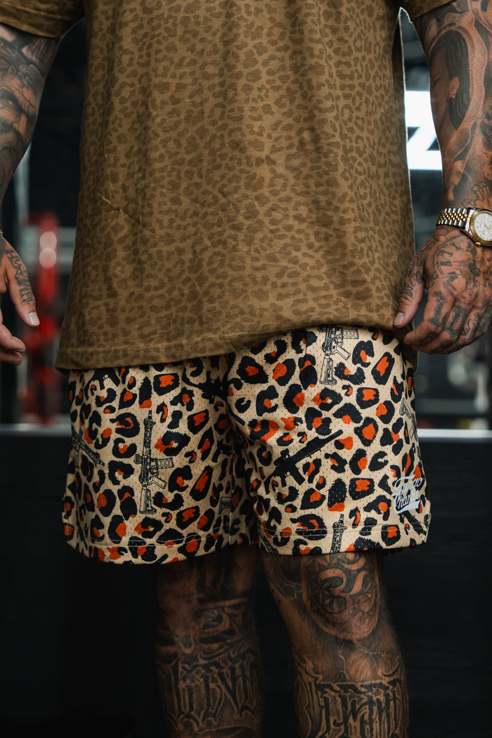 A tattooed individual wears Tango Charlie Apparel Men's Mesh Lifestyle Shorts | 5.5"- Loaded Leopard with a brown leopard print shirt and a gold watch, standing against a dark, blurred background.