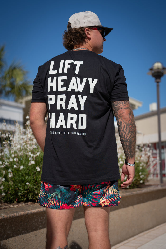 A person in a black "LIFT HEAVY PRAY HARD" shirt wears Tango Charlie Apparel's Men's Mesh Lifestyle Shorts | 5.5"- Tropical Assassin, standing outdoors in a cap and sunglasses, with plants and a building in the background.