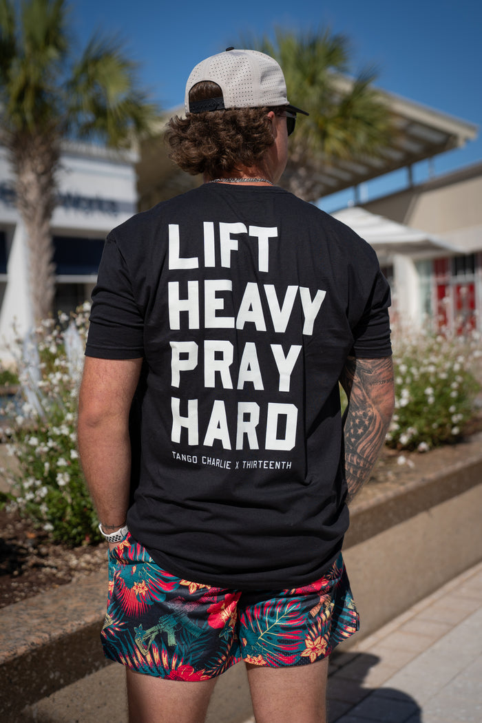 A man in a black LIFT HEAVY PRAY HARD tee, white cap, and sunglasses showcases tattooed arms and Tango Charlie Apparel’s Men's Mesh Lifestyle Shorts | 5.5"- Tropical Assassin; palm trees and a building highlight his bold style.
