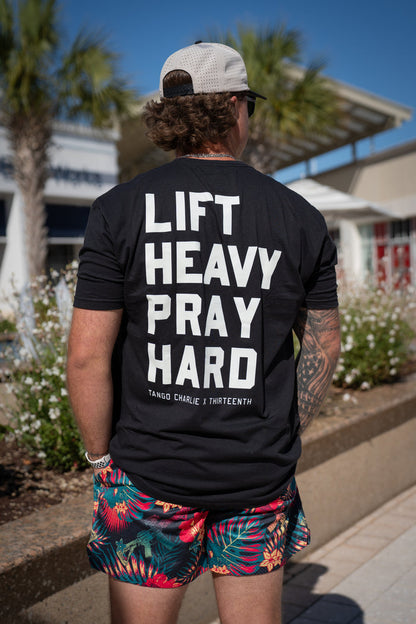 A man in a black LIFT HEAVY PRAY HARD tee, white cap, and sunglasses showcases tattooed arms and Tango Charlie Apparel’s Men's Mesh Lifestyle Shorts | 5.5"- Tropical Assassin; palm trees and a building highlight his bold style.
