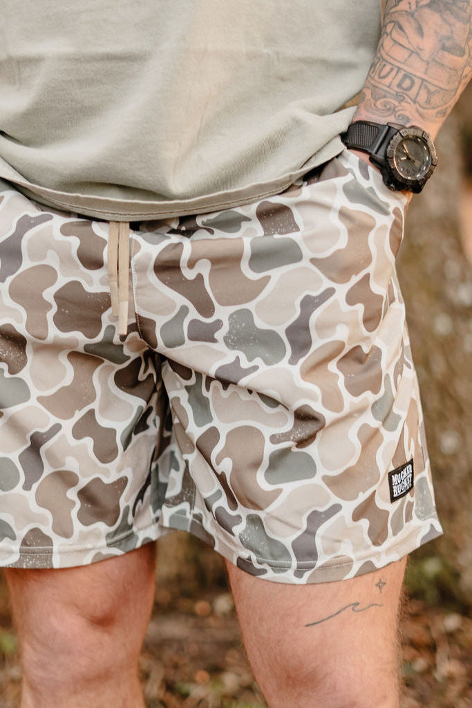 Donning Mens Performance Athletic Shorts by Mucker Bucket and a beige t-shirt, the person stands outdoors with a left-arm tattoo and black watch. Amid the peaceful treescape, they effortlessly blend into nature like a discreet duck.