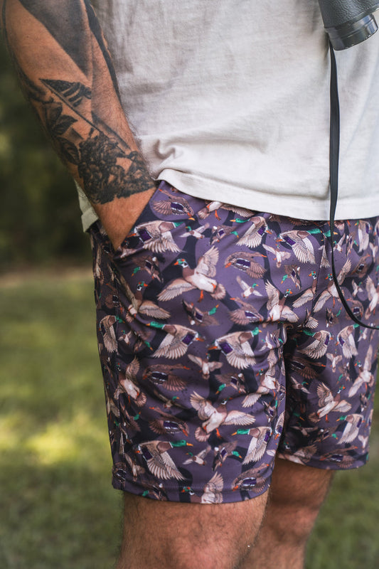 A person stands outdoors in Mucker Bucket Men's Performance Athletic Shorts | 5.5"- Duck Wild—purple shorts with flying ducks—paired with a white T-shirt. Their tattooed arm and camera strap are visible, with grass and trees in the background.
