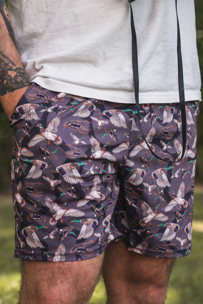 A tattooed individual stands outdoors in a white t-shirt and Mucker Bucket Men's Performance Athletic Shorts | 5.5" – Duck Wild, featuring a colorful bird pattern, with binoculars in hand amid a serene, green backdrop.