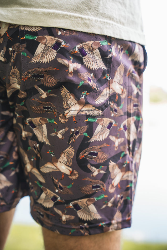 Close-up of a person wearing Mucker Bucket Men's Performance Athletic Shorts | 5.5"- Duck Wild, featuring vibrant duck illustrations on dark fabric. The blurred background highlights the shorts’ stylish and comfortable design.