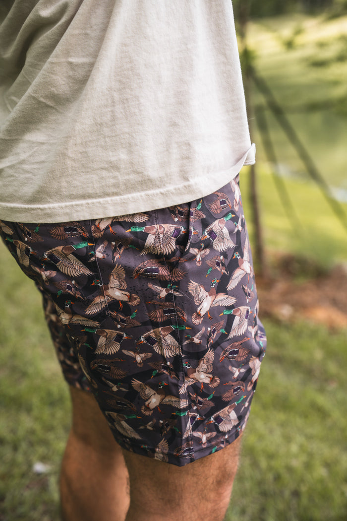 A person outdoors in a white t-shirt and Mucker Bucket Men's Performance Athletic Shorts | 5.5"- Duck Wild, featuring a colorful duck pattern, with grass and blurred trees in the background.