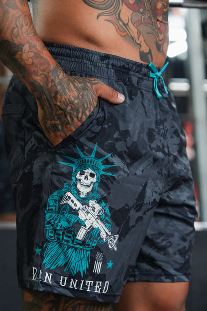 A tattooed, shirtless man stands indoors with one hand in the pocket of Born United Private Label Men's Performance Hybrid Shorts 5.5" | Liberty or Death, featuring quick-drying fabric and a skeleton Statue of Liberty design above "BIN UNITED.