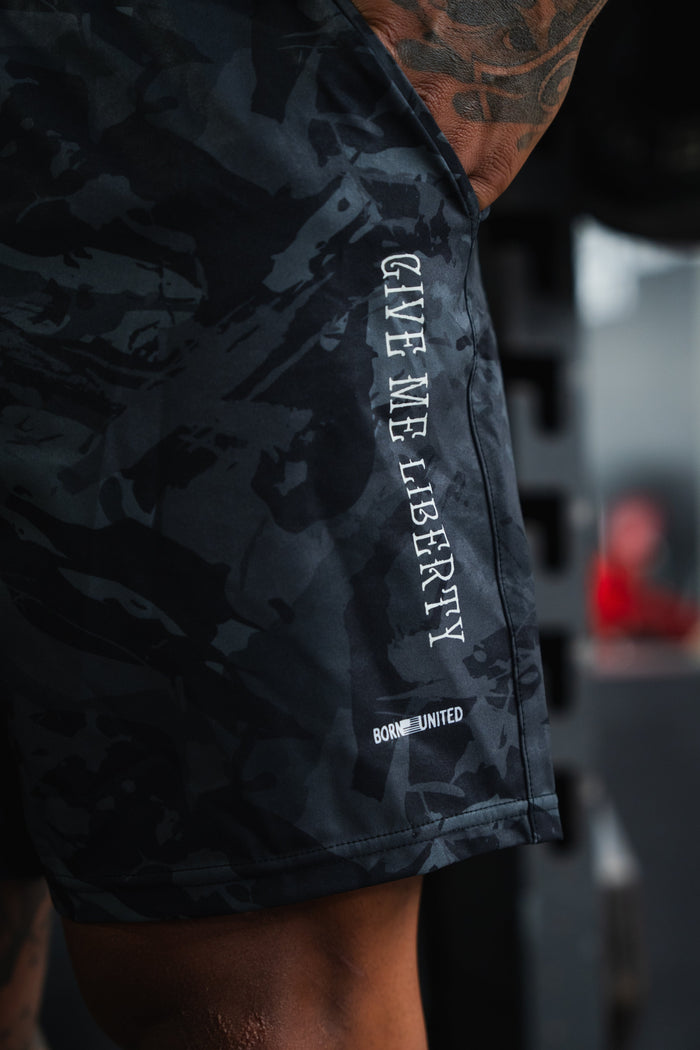 A close-up of Born United Private Label Men's Performance Hybrid Shorts 5.5" | Liberty or Death in a camo print, featuring "GIVE ME LIBERTY" printed vertically in white—quick-drying fabric and bold patriotic design. Tattooed arm and thigh partially visible.