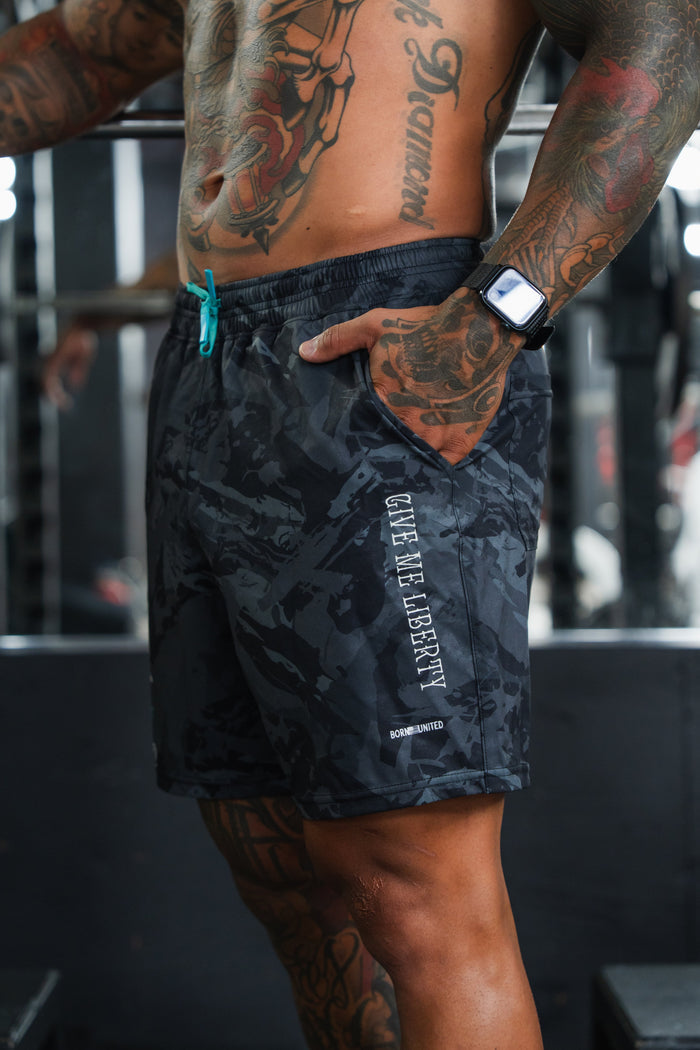 A muscular, tattooed person stands in a gym with a hand in the pocket, wearing Born United Private Label Men's Performance Hybrid Shorts 5.5" | Liberty or Death—quick-drying shorts featuring the patriotic GIVE ME LIBERTY design on the side.