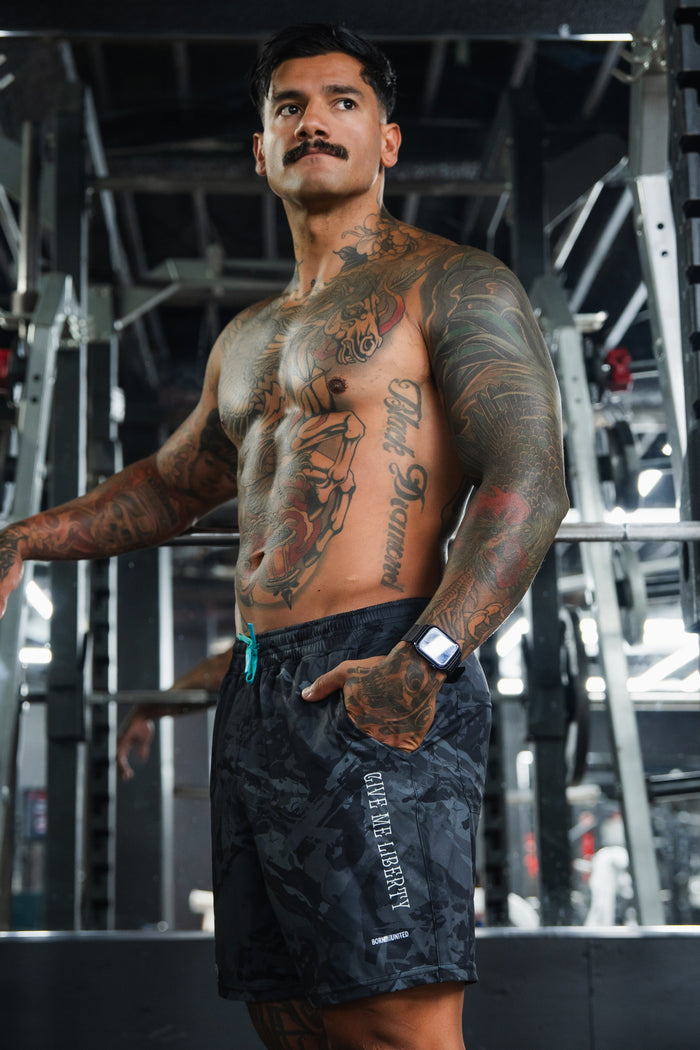 A muscular, tattooed man stands shirtless in a gym wearing Born United Private Label Men's Performance Hybrid Shorts 5.5" | Liberty or Death, featuring a patriotic design and quick-drying fabric, with gym equipment and mirrors in the background.