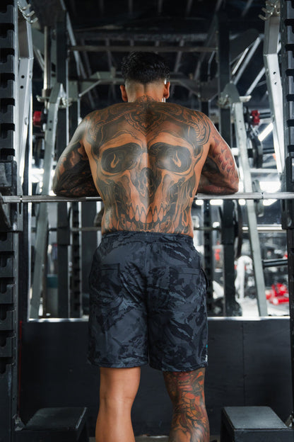 A person with a large skull tattoo on their back stands in a gym, facing equipment and preparing to lift a barbell while wearing Born United Private Label Men's Performance Hybrid Shorts 5.5" | Liberty or Death.