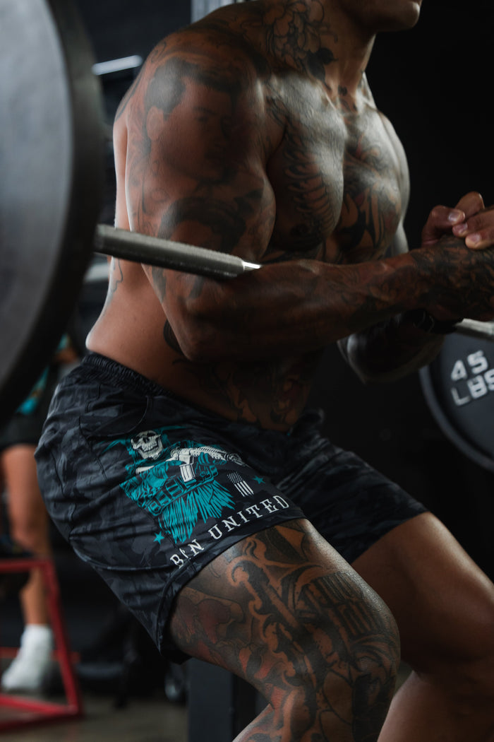 A muscular, tattooed person is squatting with a barbell at the gym, wearing Born United Private Label Men's Performance Hybrid Shorts 5.5" | Liberty or Death, featuring a quick-drying blue and white patriotic design.