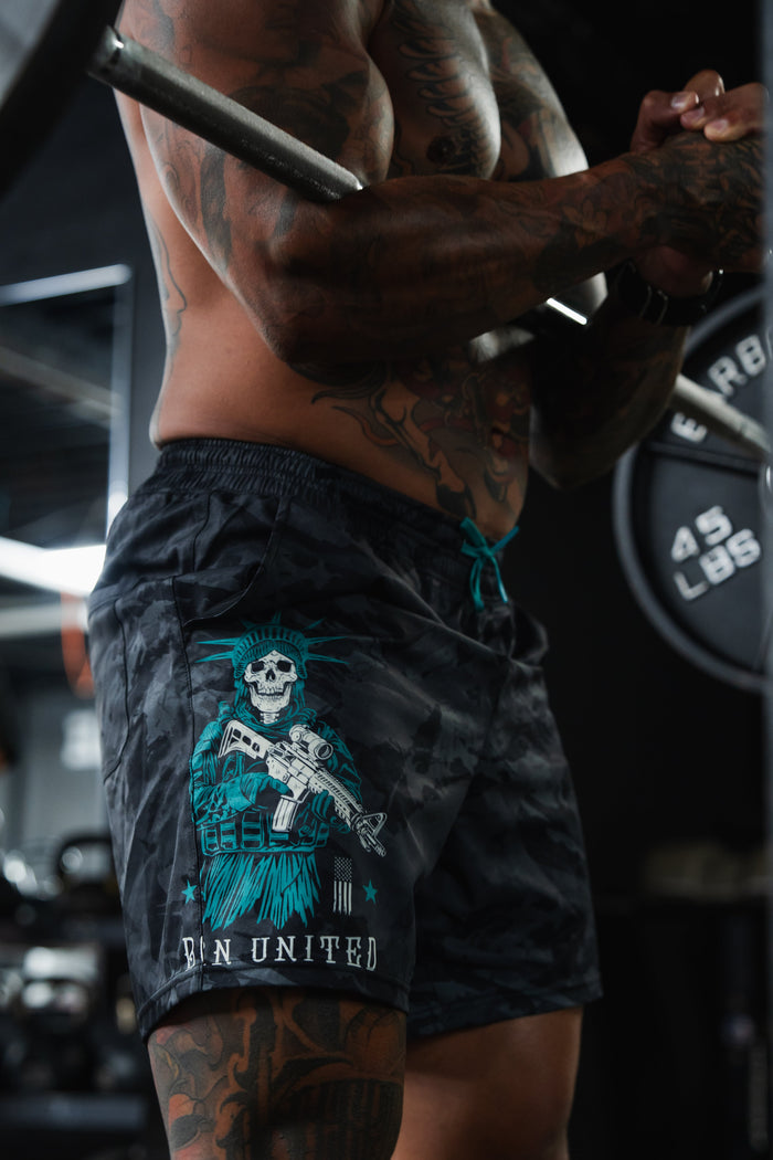 A muscular, tattooed person lifts a barbell in the gym wearing Born United Private Label Men's Performance Hybrid Shorts 5.5" | Liberty or Death, featuring a patriotic skeleton-in-crown design with “UN UNITED” text and quick-drying fabric.