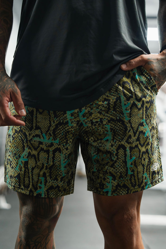 A person in a dark shirt and Tango Charlie Apparel Men's Performance Training Shorts | 5.5" - Lethal Venom stands indoors with hands on the waistband, displaying tattoos on their arms.