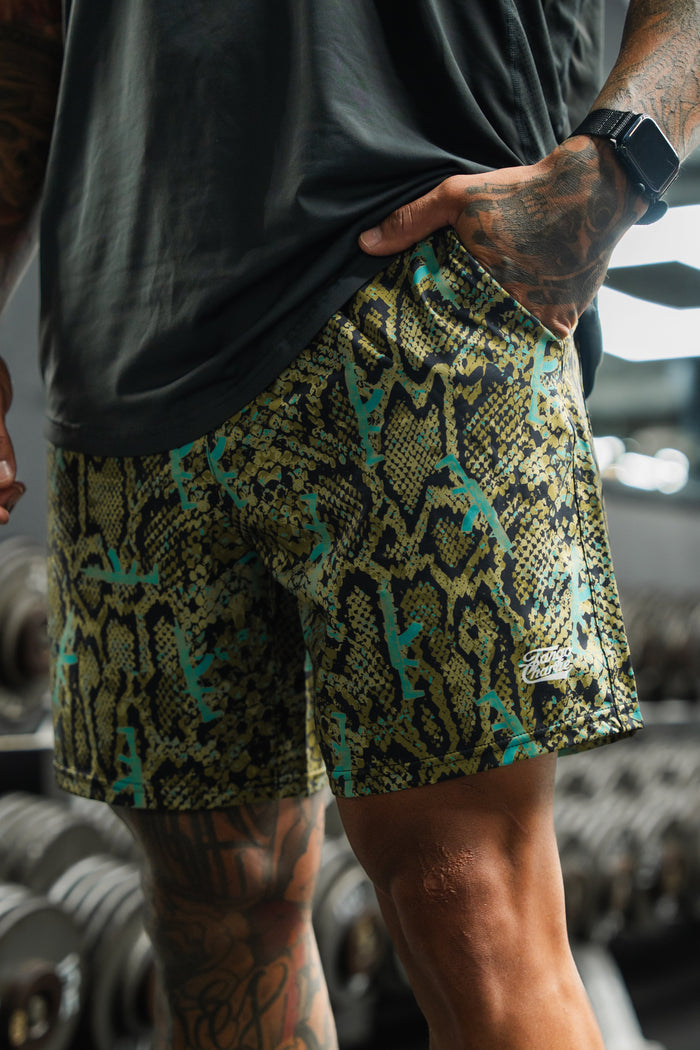 Wearing Tango Charlie Apparel Men's Performance Training Shorts | 5.5"- Lethal Venom and a black shirt, a person with tattooed arms and legs stands by gym dumbbells, sporting a smartwatch.