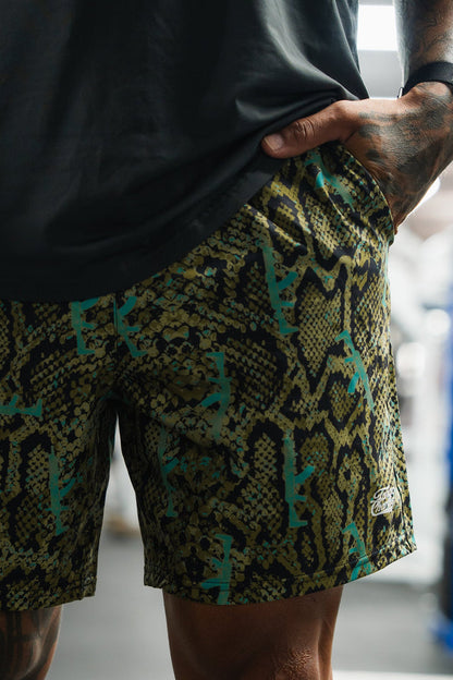 A person indoors lifts their black shirt to reveal Tango Charlie Apparel’s Men's Performance Training Shorts | 5.5"- Lethal Venom, featuring a striking green, yellow, and black snakeskin pattern.