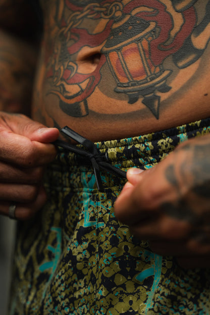 A close-up of tattooed hands tying the drawstring of Tango Charlie Apparel Men's Performance Training Shorts | 5.5"– Lethal Venom, with a detailed colorful lantern tattoo visible on the lower abdomen.
