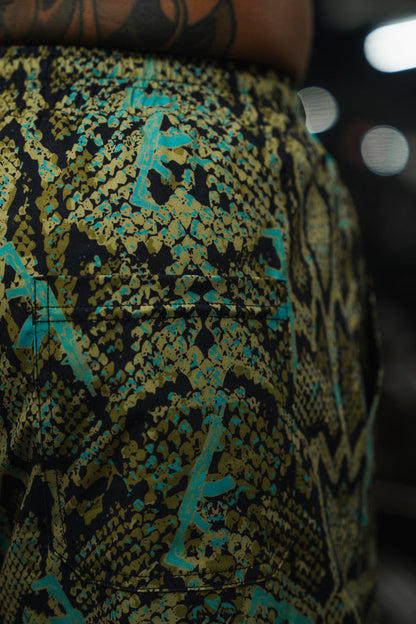 Close-up of a person wearing Tango Charlie Apparel's Men's Performance Training Shorts | 5.5"- Lethal Venom, featuring a green, yellow, and black snakeskin pattern with blue accents. A lower back tattoo edge is visible; background is blurred.