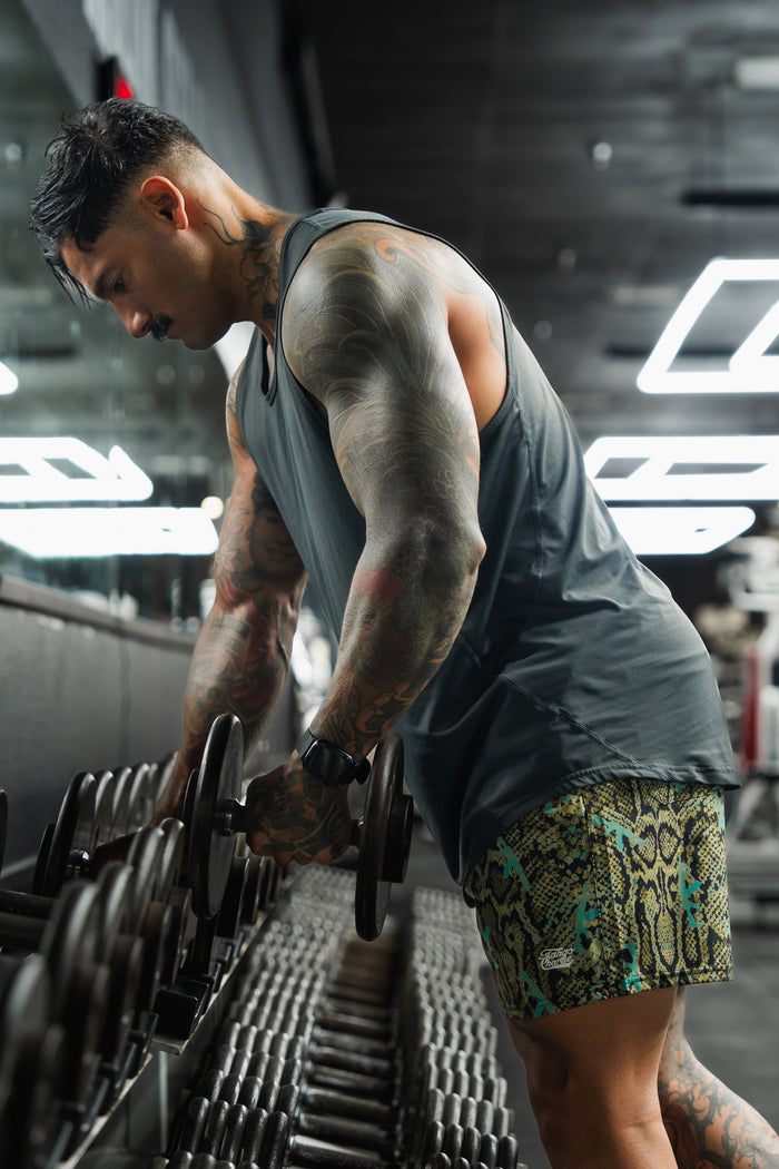 A tattooed, muscular man in a gray tank top and Tango Charlie Apparel Men's Performance Training Shorts | 5.5"- Lethal Venom picks a dumbbell from a rack in a modern, mirror-walled gym.