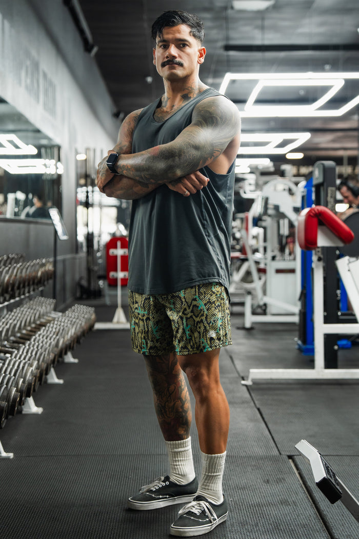 A muscular man with a mustache and tattoos stands in a gym wearing a sleeveless shirt, Tango Charlie Apparel Men's Performance Training Shorts | 5.5" - Lethal Venom, white socks, and sneakers. Gym equipment and mirrors are visible behind him.