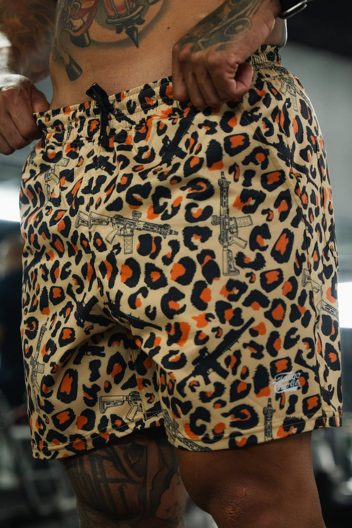 A person adjusts their Tango Charlie Apparel Men's Performance Training Shorts | 5.5"- Loaded Leopard, featuring a bold leopard print and rifle graphics, while standing in a gym with exercise equipment blurred in the background.