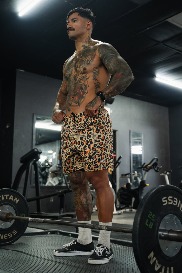A muscular, tattooed man stands shirtless in a gym, wearing Tango Charlie Apparel Men's Performance Training Shorts | 5.5" - Loaded Leopard, paired with white socks and black sneakers. He poses by a barbell amid mirrors and workout equipment.
